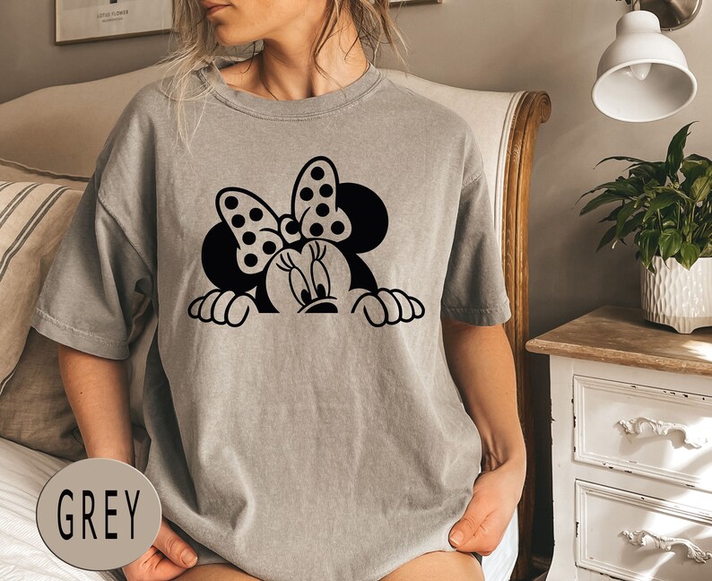 K&ouml;nnte beinhalten: Ein graues T-Shirt mit einem schwarz-wei&szlig;en Cartoon-Bild von Minnie Mouse, die &uuml;ber den oberen Rand schaut. Das Bild enth&auml;lt Minnies charakteristisches Band mit Punkten.