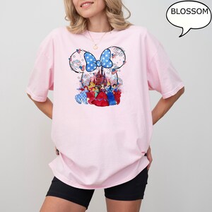 Puede incluir: Camiseta rosa claro con un dise&ntilde;o patri&oacute;tico de Minnie Mouse. El dise&ntilde;o incluye un lazo azul con estrellas blancas, fuegos artificiales y un castillo. Debajo del castillo hay varias princesas de Disney con sus vestidos.