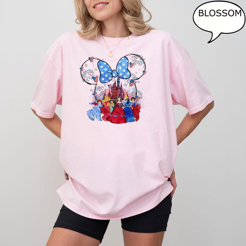 Puede incluir: Camiseta rosa claro con un dise&ntilde;o patri&oacute;tico de Minnie Mouse. El dise&ntilde;o incluye un lazo azul con estrellas blancas, fuegos artificiales y un castillo. Debajo del castillo hay varias princesas de Disney con sus vestidos.