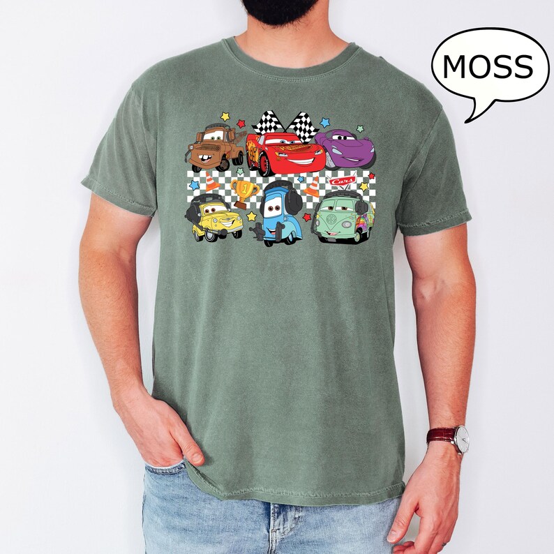 May include: A green t-shirt with a checkered pattern and a graphic of cartoon cars from the movie Cars. The cars are in a line and include Lightning McQueen, Mater, and others. The text "MOSS" is in a speech bubble above the graphic.