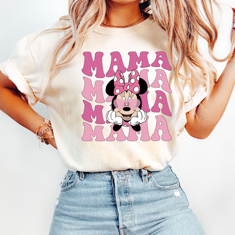Puede incluir: Camiseta blanca con un gr&aacute;fico rosa y blanco de Minnie Mouse con gafas de sol en forma de coraz&oacute;n y el texto "MAMA" repetido cuatro veces.