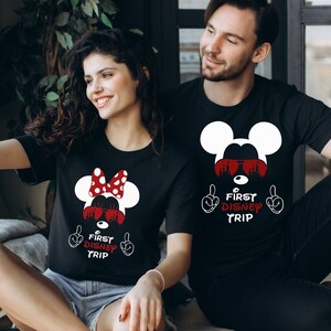 May include: A couple wearing black t-shirts with a white Mickey Mouse design and the text "First Disney Trip" in red. The woman's shirt features a red bow on Mickey Mouse's head.