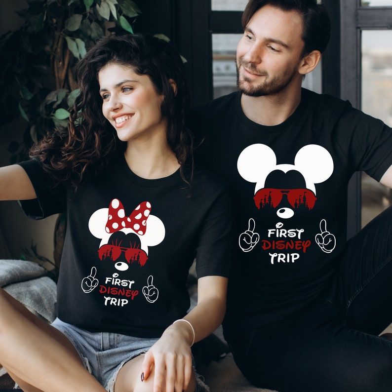 May include: A couple wearing black t-shirts with a white Mickey Mouse design and the text "First Disney Trip" in red. The woman's shirt features a red bow on Mickey Mouse's head.