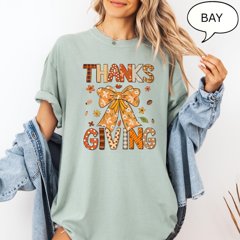 May include: A light green t-shirt with a Thanksgiving graphic. The graphic features the words "THANKS GIVING" in a mix of patterns and colors, including orange, brown, and white. A large bow and floral elements are also included in the design.