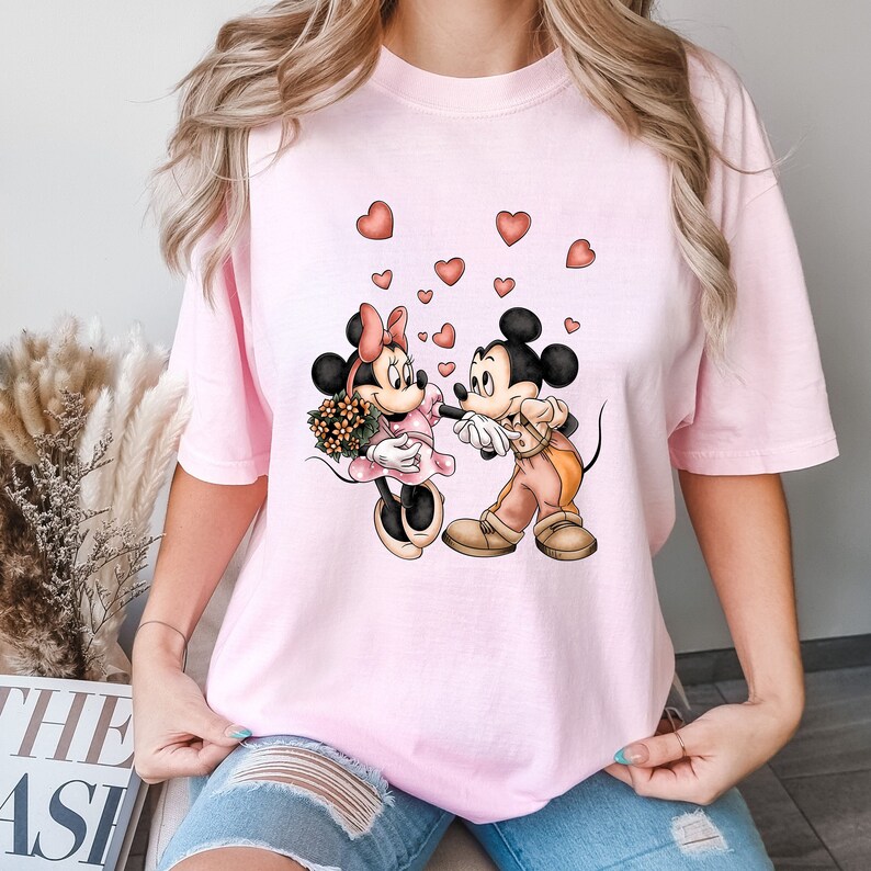 May include: Pink t-shirt with a graphic of Mickey Mouse and Minnie Mouse holding hands with hearts floating around them.