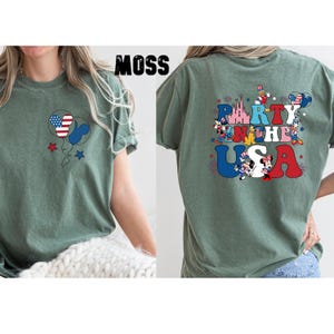 K&ouml;nnte beinhalten: Moosgr&uuml;nes T-Shirt mit patriotischem Design. Die Vorderseite zeigt ein Ballondesign in den Farben der amerikanischen Flagge. Auf der R&uuml;ckseite des Shirts steht "Party in the USA" mit Zeichentrickfiguren und patriotischen Bildern.