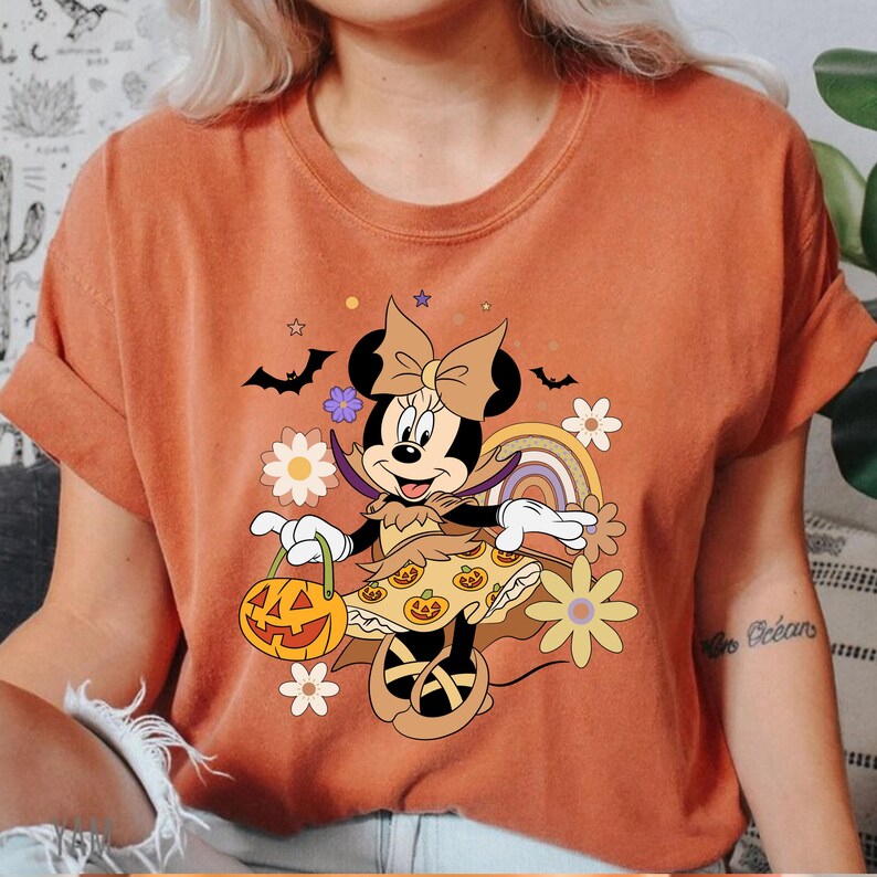 Puede incluir: Camiseta naranja con un dise&ntilde;o de Minnie Mouse de Halloween. El gr&aacute;fico muestra a Minnie disfrazada, sosteniendo una linterna de calabaza, con flores, murci&eacute;lagos y un arco&iacute;ris. La camiseta tiene cuello redondo y mangas cortas.