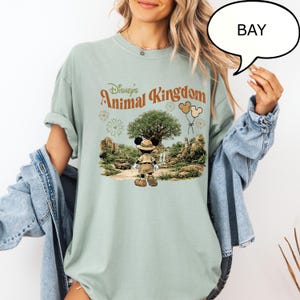 May include: A light green t-shirt featuring the text "Disney's Animal Kingdom" in a vintage style. The graphic includes Mickey Mouse in safari attire, a large tree, and balloons. The shirt has rolled-up sleeves.