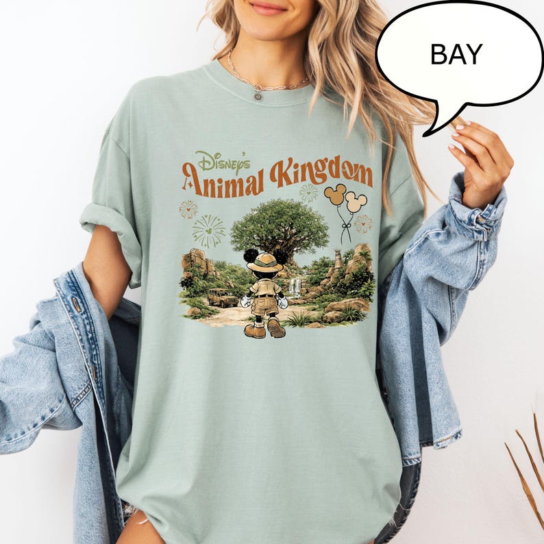 May include: A light green t-shirt featuring the text "Disney's Animal Kingdom" in a vintage style. The graphic includes Mickey Mouse in safari attire, a large tree, and balloons. The shirt has rolled-up sleeves.