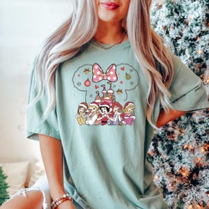 Puede incluir: Una camiseta verde claro con un gr&aacute;fico navide&ntilde;o que presenta un lazo rosa, un castillo y siete princesas Disney con gorros de Pap&aacute; Noel. Las princesas est&aacute;n de pie frente al castillo, que est&aacute; decorado con luces navide&ntilde;as y adornos.