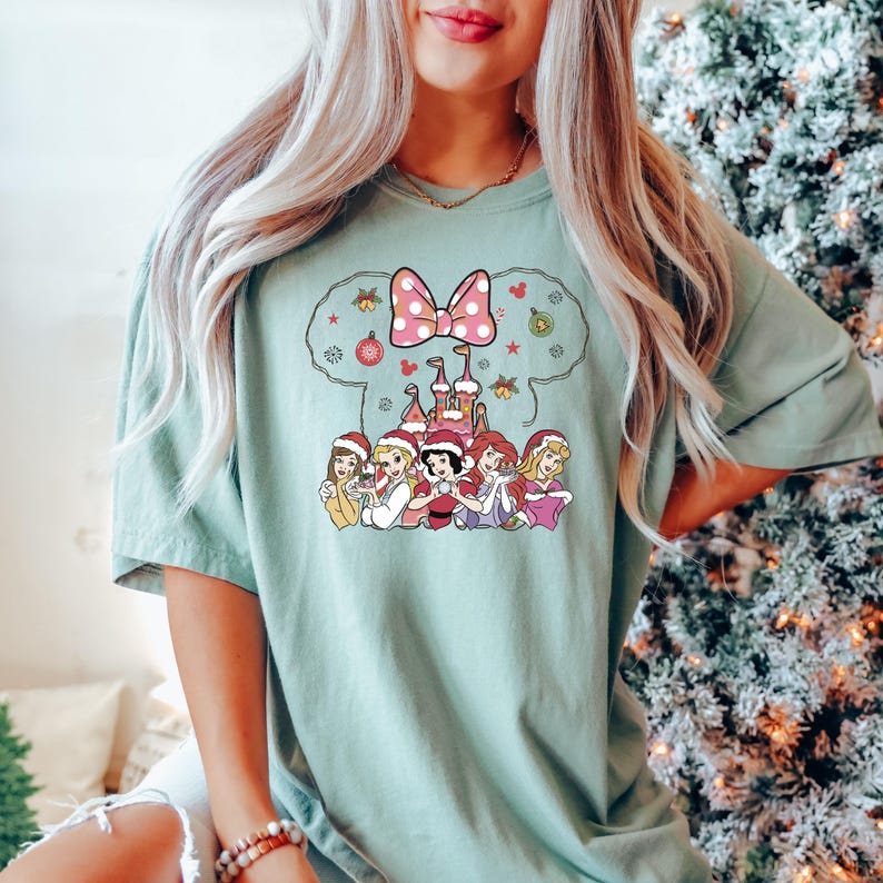 Puede incluir: Una camiseta verde claro con un gr&aacute;fico navide&ntilde;o que presenta un lazo rosa, un castillo y siete princesas Disney con gorros de Pap&aacute; Noel. Las princesas est&aacute;n de pie frente al castillo, que est&aacute; decorado con luces navide&ntilde;as y adornos.