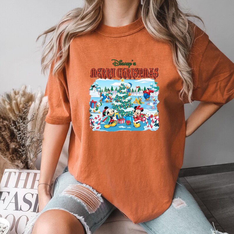 May include: Orange t-shirt with a Christmas-themed graphic featuring Mickey Mouse, Minnie Mouse, Donald Duck, and Goofy. The graphic says "Disney's Merry Christmas".
