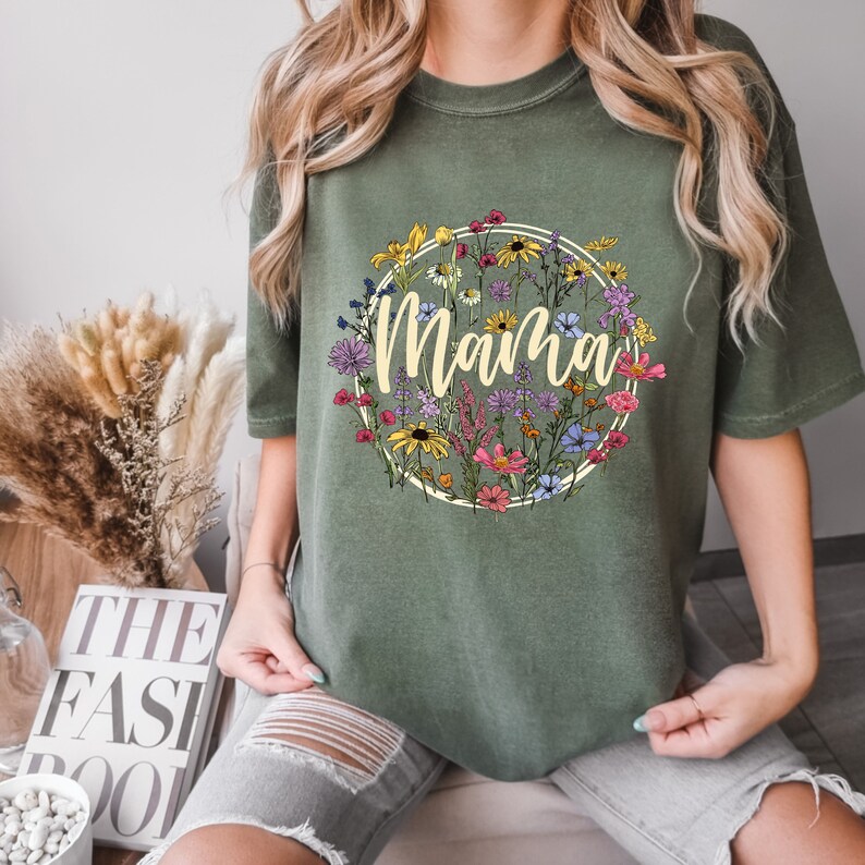 Peut inclure: Un t-shirt vert avec un motif circulaire floral et le mot "Mama" en police cr&egrave;me.