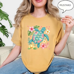 May include: Mustard-colored t-shirt featuring a turquoise sea turtle with a floral design. The turtle is surrounded by pink and yellow flowers and green leaves. The word "MUSTARD" is in a speech bubble.