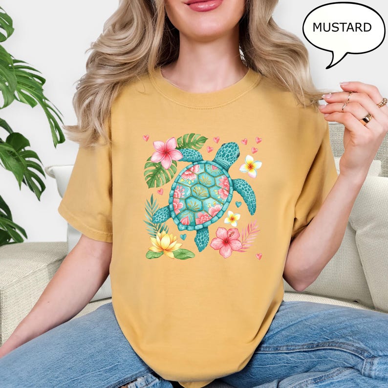 May include: Mustard-colored t-shirt featuring a turquoise sea turtle with a floral design. The turtle is surrounded by pink and yellow flowers and green leaves. The word "MUSTARD" is in a speech bubble.