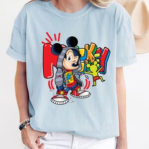 Pode incluir: Camiseta azul claro com um gr&aacute;fico colorido do Mickey Mouse vestindo uma jaqueta vermelha e azul, cal&ccedil;as vermelhas e t&ecirc;nis preto e branco. O gr&aacute;fico tamb&eacute;m inclui a palavra "Mickey" em um estilo de graffiti colorido.