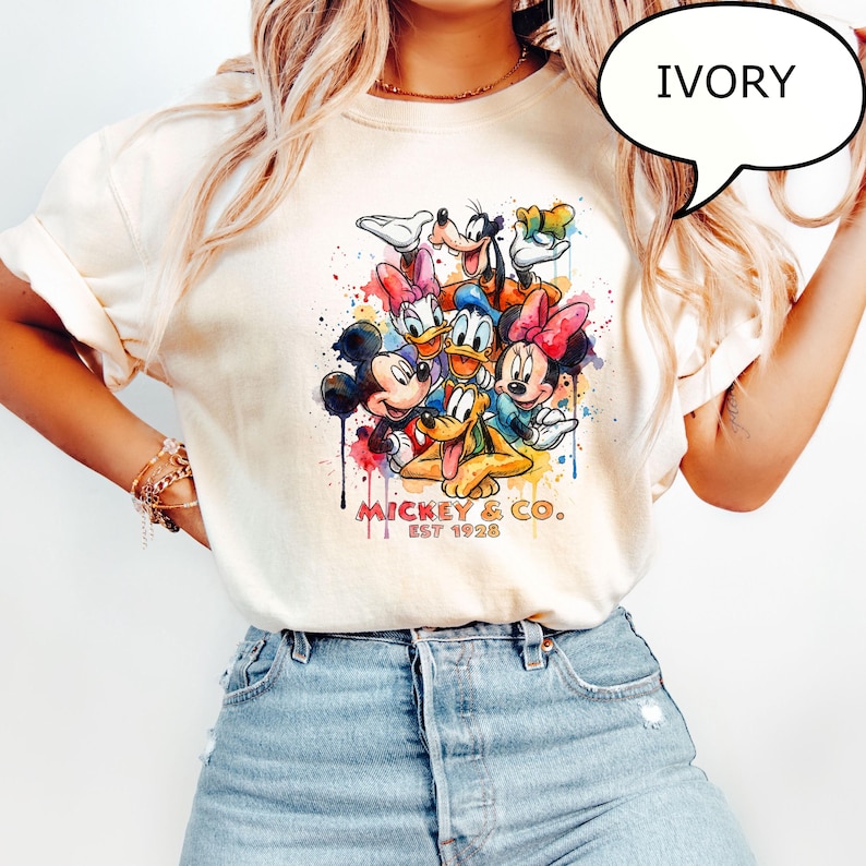 May include: Ivory-colored t-shirt featuring a colorful watercolor design of Mickey Mouse, Minnie Mouse, Donald Duck, Daisy Duck, Goofy, and Pluto. The shirt includes the text "Mickey & Co. EST 1928".
