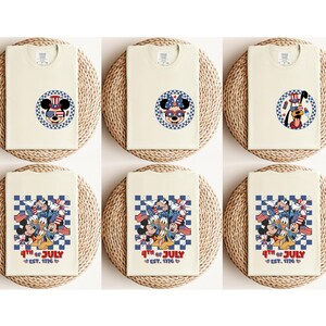 Pode incluir: T-shirts creme com designs patri&oacute;ticos com Mickey Mouse, Minnie Mouse e Pluto. Os designs incluem elementos vermelhos, brancos e azuis e o texto "4th of July EST. 1776". As camisas s&atilde;o exibidas em esteiras tecidas.