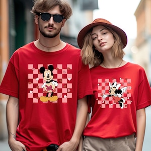 Puede incluir: Camisetas rojas con dise&ntilde;os de Mickey y Minnie Mouse. Mickey Mouse sostiene una caja de regalo en forma de coraz&oacute;n sobre un fondo a cuadros rosa y blanco. Minnie Mouse se representa como Cupido con arco y flecha.