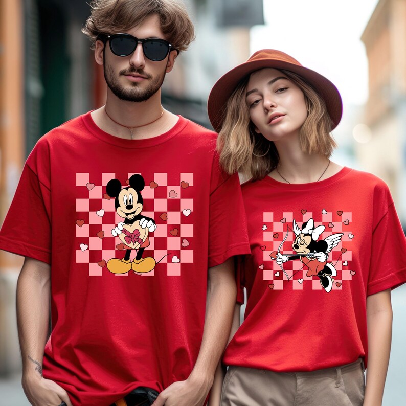 Puede incluir: Camisetas rojas con dise&ntilde;os de Mickey y Minnie Mouse. Mickey Mouse sostiene una caja de regalo en forma de coraz&oacute;n sobre un fondo a cuadros rosa y blanco. Minnie Mouse se representa como Cupido con arco y flecha.