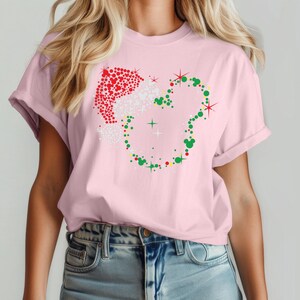 Puede incluir: Una camiseta rosa claro con un dise&ntilde;o navide&ntilde;o. El dise&ntilde;o incluye un gorro de Pap&aacute; Noel hecho de puntos rojos y blancos, y una forma de cabeza de Mickey Mouse hecha de puntos verdes. Es una prenda de vestir informal.