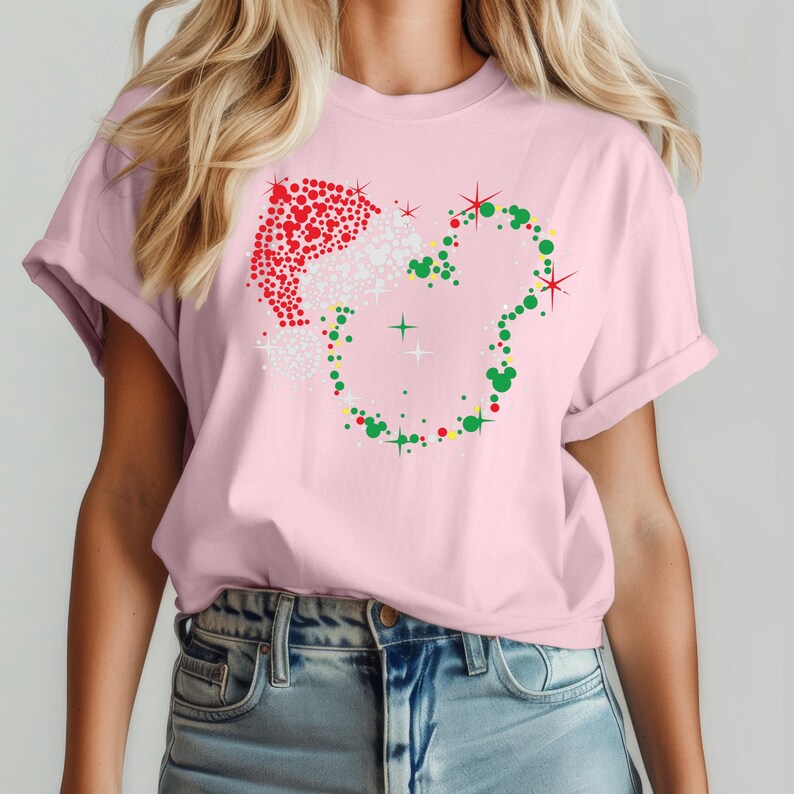 Puede incluir: Una camiseta rosa claro con un dise&ntilde;o navide&ntilde;o. El dise&ntilde;o incluye un gorro de Pap&aacute; Noel hecho de puntos rojos y blancos, y una forma de cabeza de Mickey Mouse hecha de puntos verdes. Es una prenda de vestir informal.