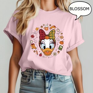 May include: A light pink t-shirt featuring a cartoon graphic of Daisy Duck with a fall-themed headband and surrounded by autumn elements. The word "BLOSSOM" is in a speech bubble. The shirt is a casual wear item.