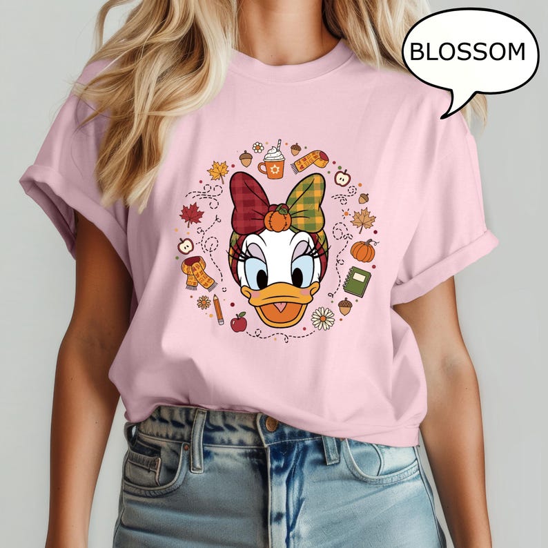 May include: A light pink t-shirt featuring a cartoon graphic of Daisy Duck with a fall-themed headband and surrounded by autumn elements. The word "BLOSSOM" is in a speech bubble. The shirt is a casual wear item.