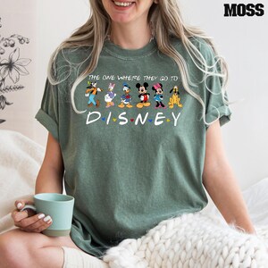 May include: Green t-shirt with the text "THE ONE WHERE THEY GO TO DISNEY" and the word "DISNEY" below it. The shirt features cartoon characters including Goofy, Daisy Duck, Donald Duck, Mickey Mouse, Minnie Mouse, and Pluto.
