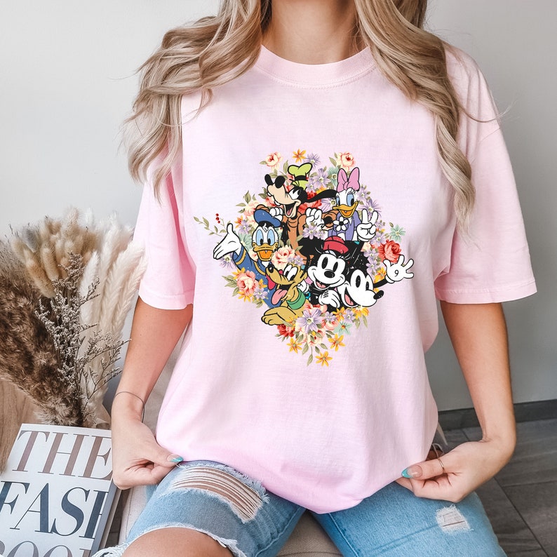 May include: Pink t-shirt featuring a graphic print of Mickey Mouse, Minnie Mouse, Donald Duck, Goofy, Pluto, and Daisy Duck surrounded by flowers.