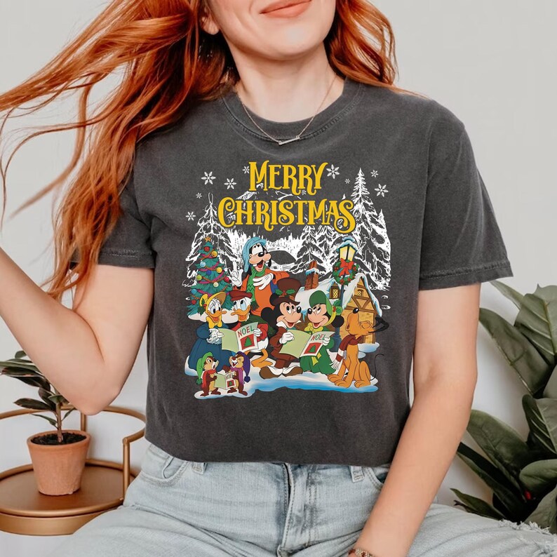 Puede incluir: Una camiseta gris oscuro con un dise&ntilde;o navide&ntilde;o vintage con Mickey Mouse, Minnie Mouse, Donald Duck, Goofy, Pluto y otros personajes de Disney cantando villancicos. El texto "Merry Christmas" est&aacute; impreso en amarillo en la camiseta.