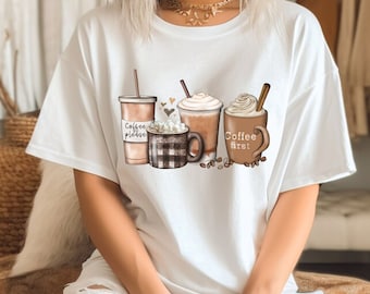 Puede incluir: Camiseta blanca con un gráfico de cuatro bebidas de café. Las bebidas son un vaso de café para llevar, una taza con malvaviscos, un latte y un café con crema batida. El texto en las tazas dice "Coffee please" y "Coffee first".