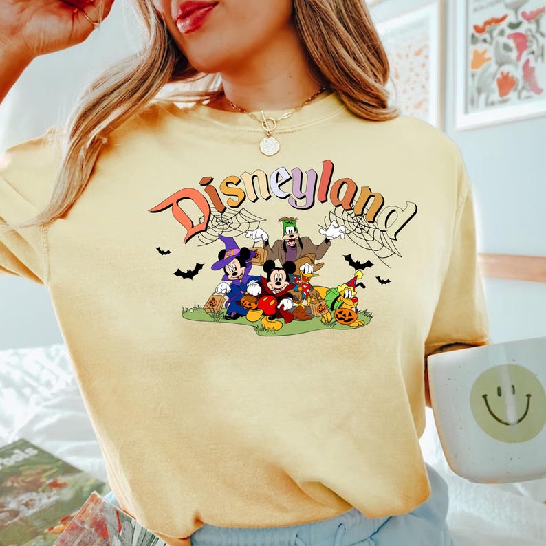 Puede incluir: Una camiseta amarilla con un estampado gr&aacute;fico de Mickey Mouse y sus amigos vestidos con disfraces de Halloween. El texto "Disneyland" est&aacute; impreso en una fuente retro sobre los personajes.