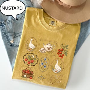 Puede incluir: Una camiseta de color amarillo mostaza con un estampado gr&aacute;fico de dos ocas, una tetera con una taza de t&eacute;, una cesta de fresas, un racimo de tomates rojos y varias ilustraciones florales. La camiseta tiene cuello redondo y etiqueta Comfort Colors.