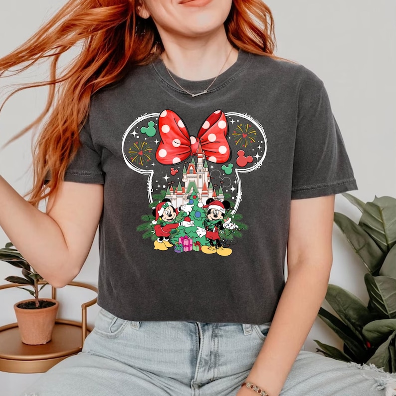 Puede incluir: Una camiseta gris oscuro recortada con un contorno blanco de la cabeza de Mickey Mouse. Dentro del contorno hay un lazo rojo y blanco con lunares, un &aacute;rbol de Navidad que Mickey y Minnie Mouse est&aacute;n decorando, y un castillo en el fondo.