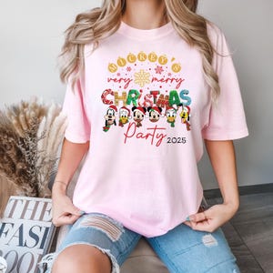 K&ouml;nnte beinhalten: Rosa T-Shirt mit einem Aufdruck von Mickey Mouse und seinen Freunden, die Weihnachten feiern. Der Text auf dem T-Shirt lautet "Mickey's Very Merry Christmas Party 2025".