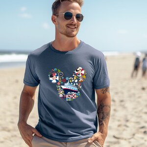 Pu&ograve; includere: T-shirt blu con un design a forma di testa di Topolino riempito di elementi nautici e dei cartoni animati. Il design include una nave da crociera, Minnie Mouse e altri personaggi. La maglietta &egrave; indossata da una persona sulla spiaggia.