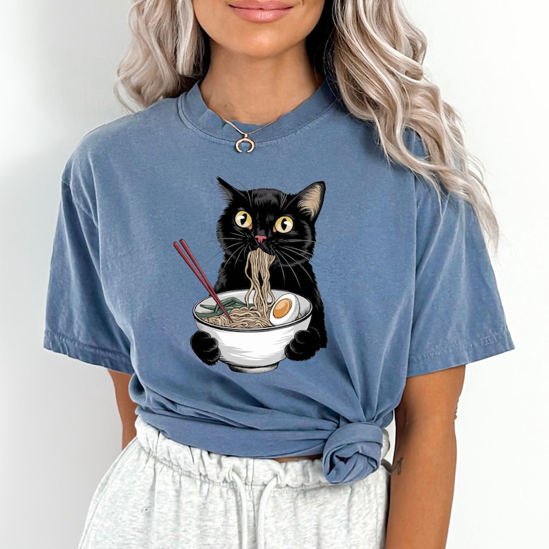 Puede incluir: Una camiseta azul con un gato negro comiendo fideos ramen de un taz&oacute;n blanco. El gato sostiene palillos en sus patas.