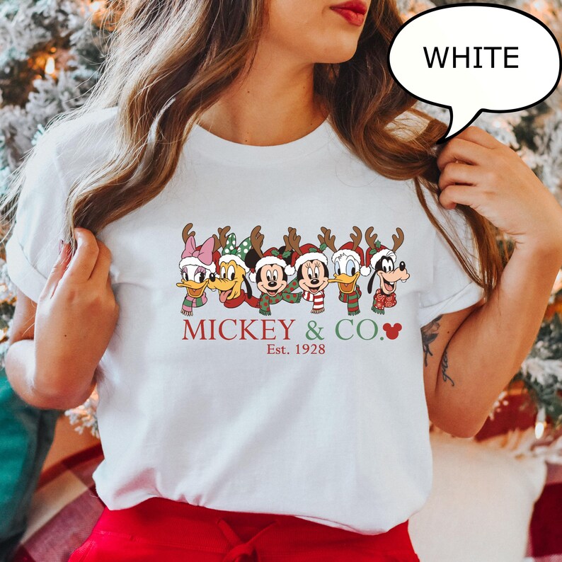 Puede incluir: Camiseta blanca con un dise&ntilde;o festivo de personajes de Disney con cuernos de reno y bufandas. El texto "Mickey & Co. Est. 1928" est&aacute; impreso en rojo, con un s&iacute;mbolo de coraz&oacute;n. Cuello redondo.