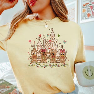 May include: A light yellow sweatshirt featuring a gingerbread castle and gingerbread characters. The gingerbread characters are decorated with red bows and candy details. A string of colorful lights and balloons are also included in the design.