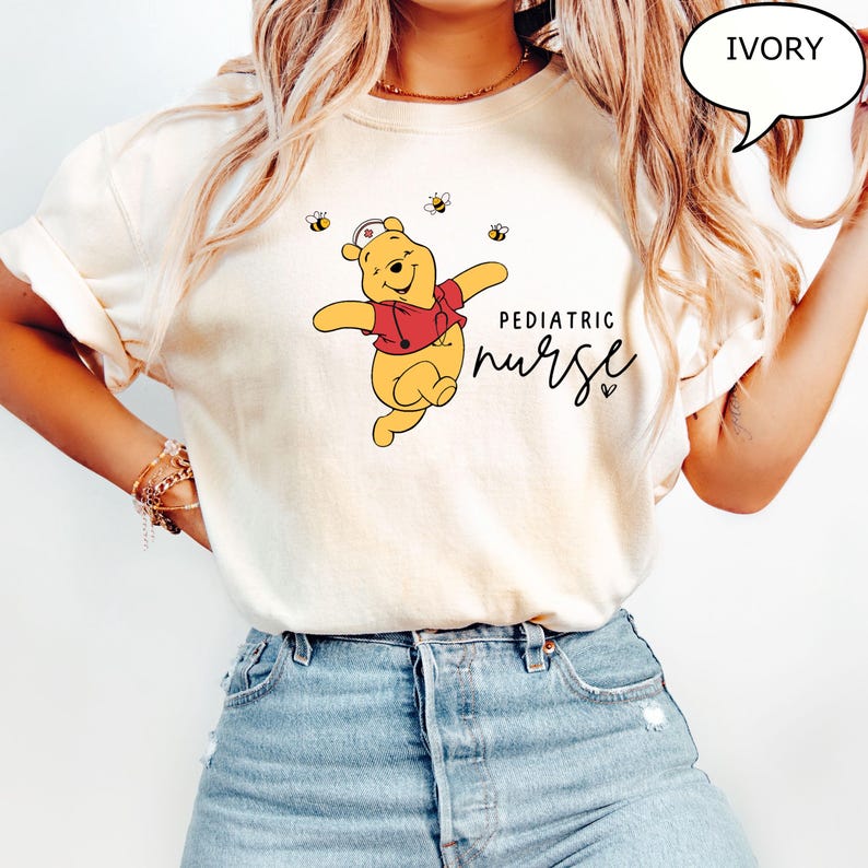 May include: Ivory-colored t-shirt featuring a cartoon Winnie the Pooh character in a red shirt and nurse's cap, surrounded by bees. The words "Pediatric Nurse" are written in black script. The shirt is paired with blue jeans.