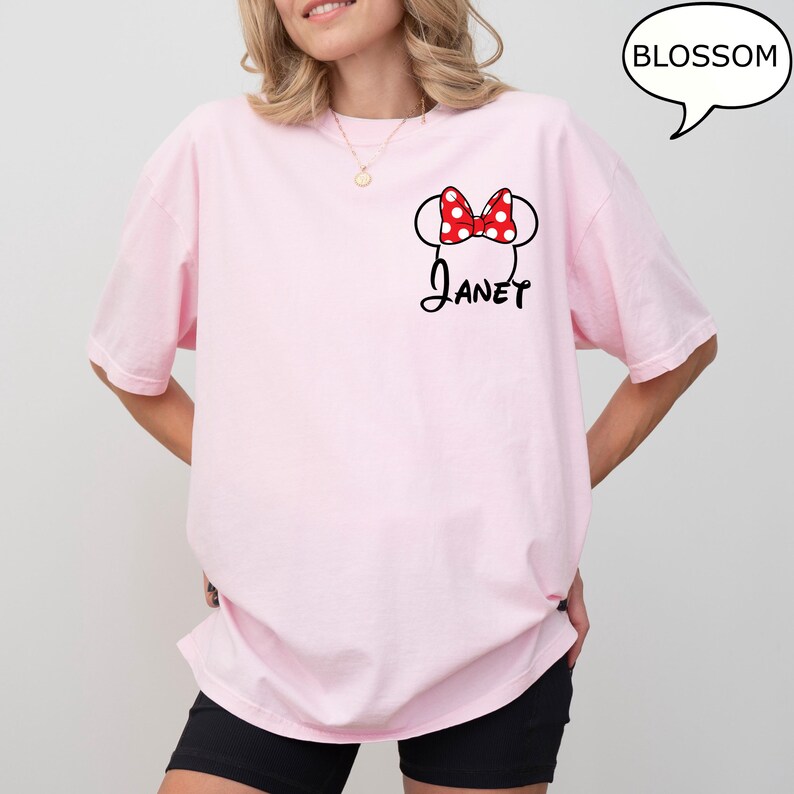 May include: A light pink t-shirt featuring a Minnie Mouse design with a red polka dot bow and the name "Janet" in black script. The shirt has short sleeves and a relaxed fit, perfect for casual wear.