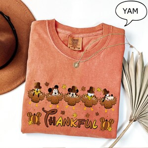May include: A coral-colored Comfort Colors t-shirt featuring a Thanksgiving-themed graphic. The design includes cartoon characters dressed as turkeys, the word "THANKFUL", and fall leaves. A gold necklace and a brown hat are also visible.