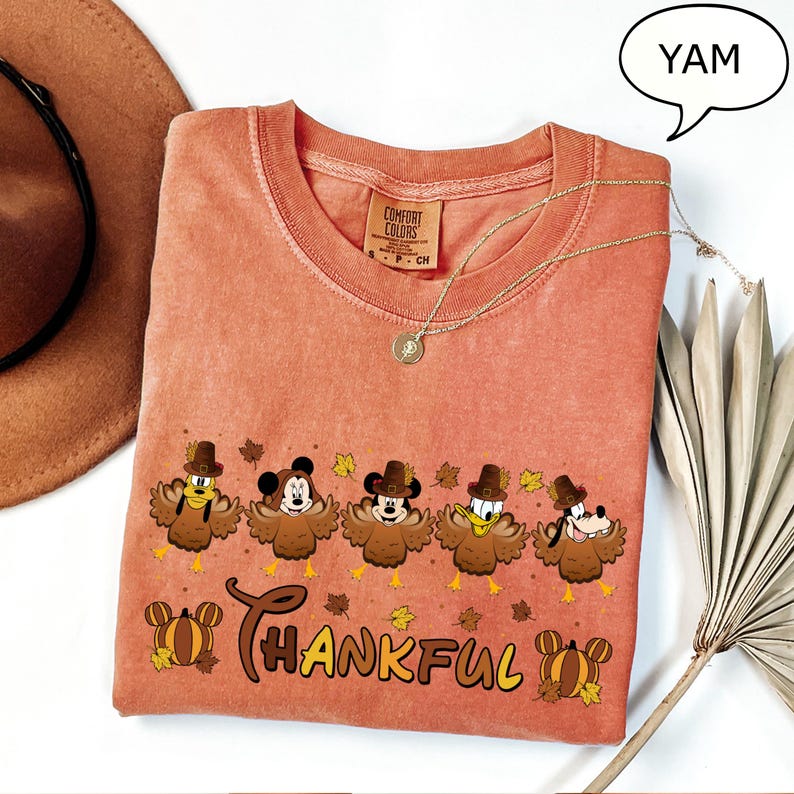 May include: A coral-colored Comfort Colors t-shirt featuring a Thanksgiving-themed graphic. The design includes cartoon characters dressed as turkeys, the word "THANKFUL", and fall leaves. A gold necklace and a brown hat are also visible.