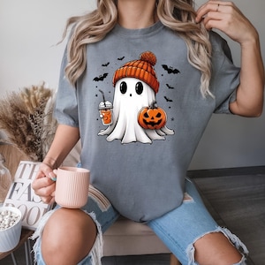 May include: A grey t-shirt featuring a cartoon ghost wearing a pumpkin-coloured hat and holding a pumpkin and a drink. The ghost has large eyes and a small mouth. There are bats and stars in the background.