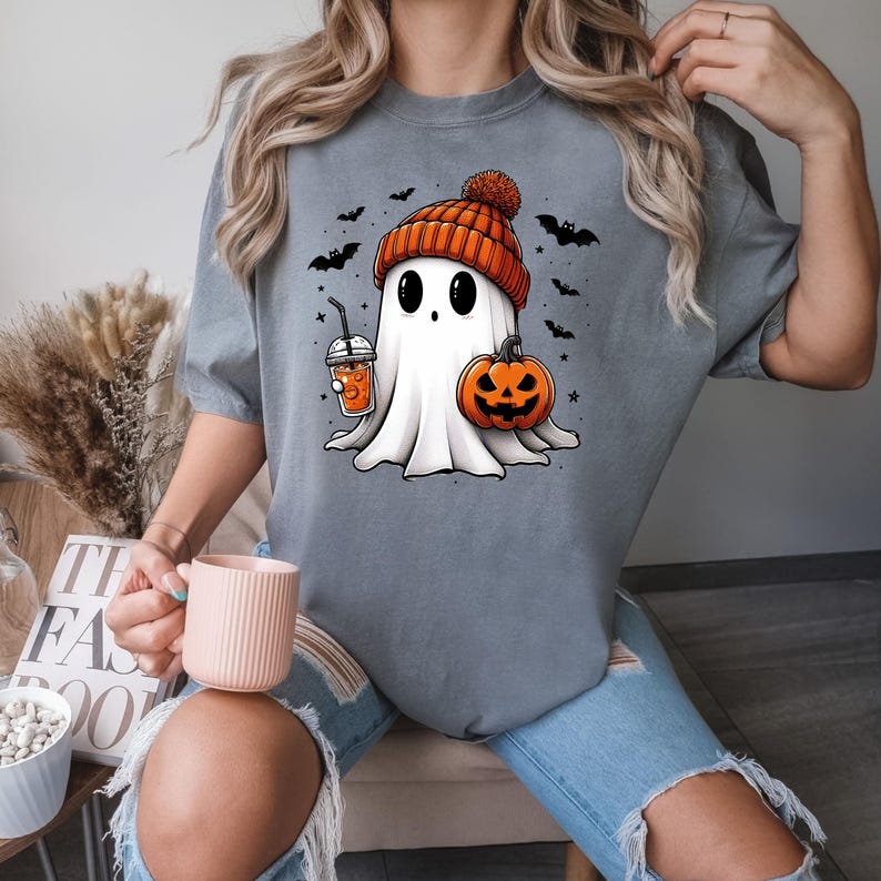 May include: A grey t-shirt featuring a cartoon ghost wearing a pumpkin-coloured hat and holding a pumpkin and a drink. The ghost has large eyes and a small mouth. There are bats and stars in the background.