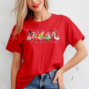 Puede incluir: Camiseta roja con un dise&ntilde;o navide&ntilde;o con cinco princesas Disney que llevan gorros de Pap&aacute; Noel y sostienen adornos. Las princesas est&aacute;n alineadas en una fila con luces de Navidad y copos de nieve detr&aacute;s de ellas.