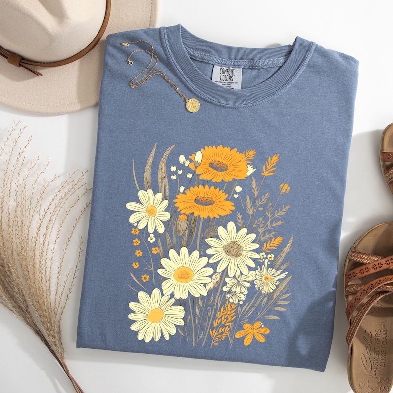 May include: A blue t-shirt featuring a floral design with yellow and white daisies and sunflowers. The shirt is styled with a gold necklace, a beige hat, and brown sandals. The shirt has the brand name "Comfort Colors" on the tag.