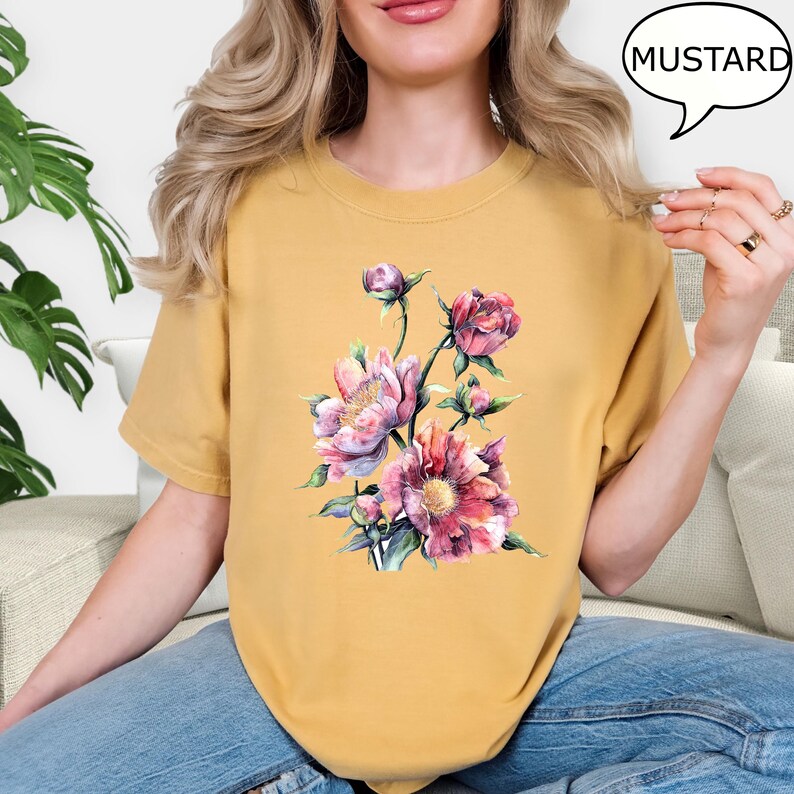 Pu&ograve; includere: T-shirt color senape con un design floreale ad acquerello nei toni del rosa e del viola. La maglietta ha girocollo e maniche corte. La parola "MUSTARD" &egrave; in una nuvoletta.