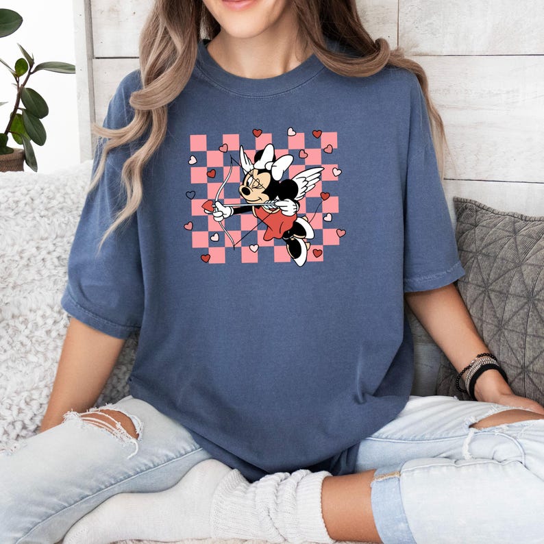 Puede incluir: Una camiseta azul con una caricatura de Minnie Mouse como Cupido, con alas y arco y flecha. El gr&aacute;fico est&aacute; sobre un fondo a cuadros rosa y blanco con detalles de coraz&oacute;n. La camiseta es de un estilo c&oacute;modo e informal.